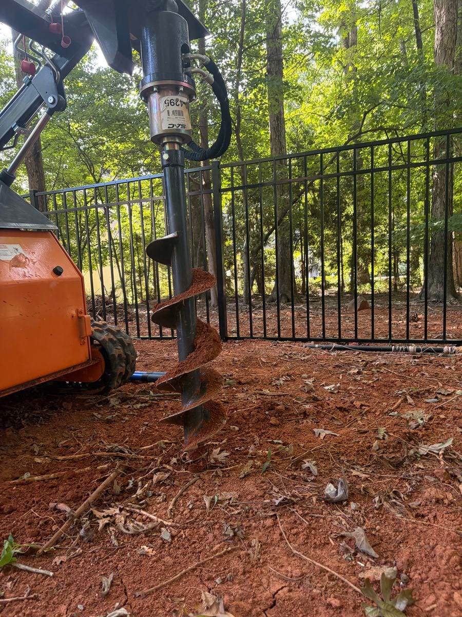 Auger drilling a post hole beside a new black aluminum fence