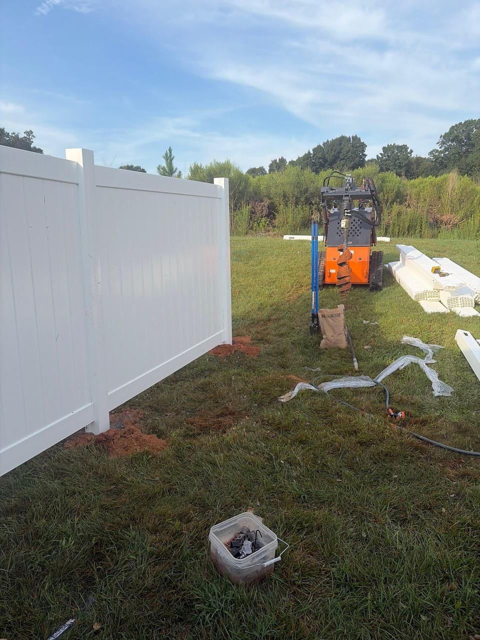 White vinyl privacy fence going in, with an auger machine and fence parts on site