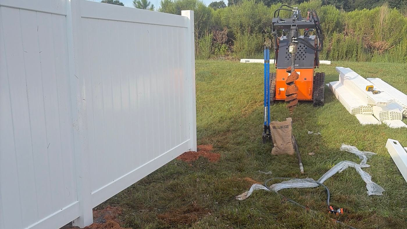 White vinyl privacy fence going in, with an auger machine and fence parts on site
